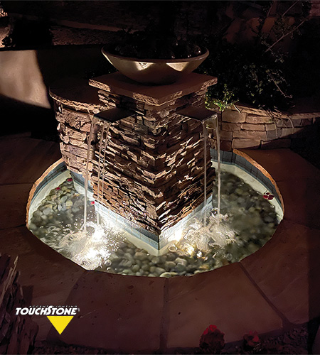 An illuminated Snail Underwater Series built with stacked stones, including a round basin on top, surrounded by a pond with pebbles and snails. Nighttime setting.