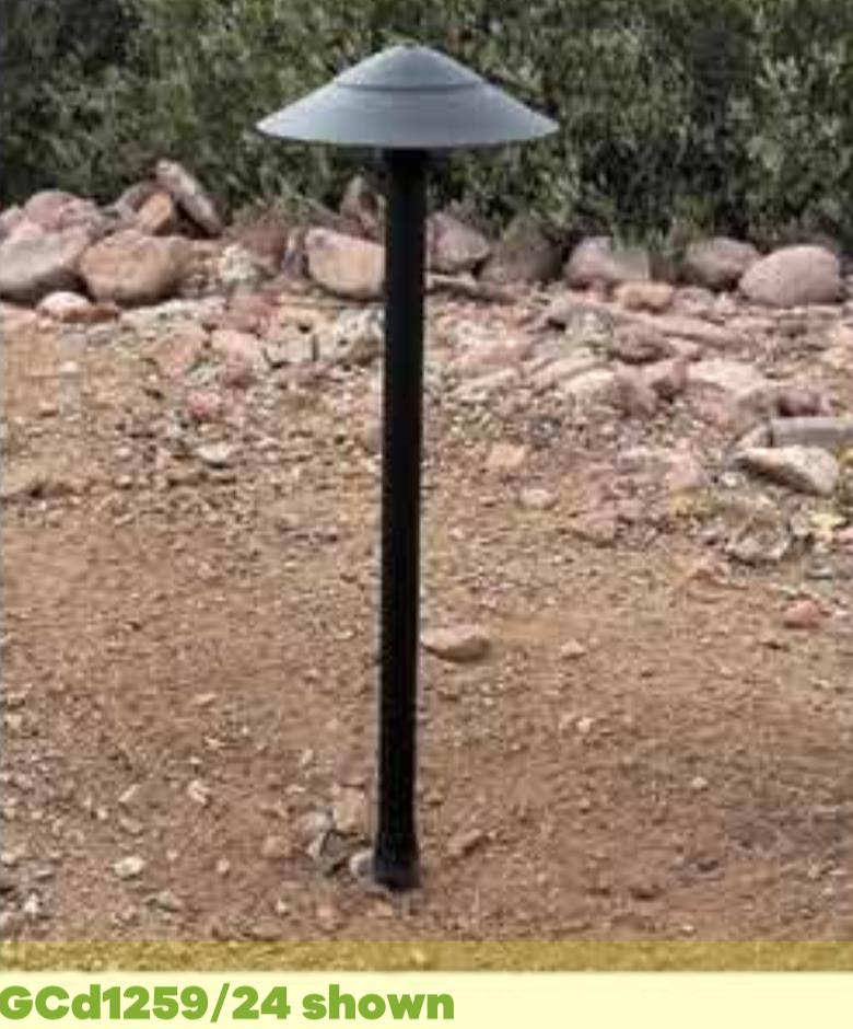 A Commander black outdoor pathway light, featuring a dome-shaped top, is set among rocks and earth, with shrubs in the background.