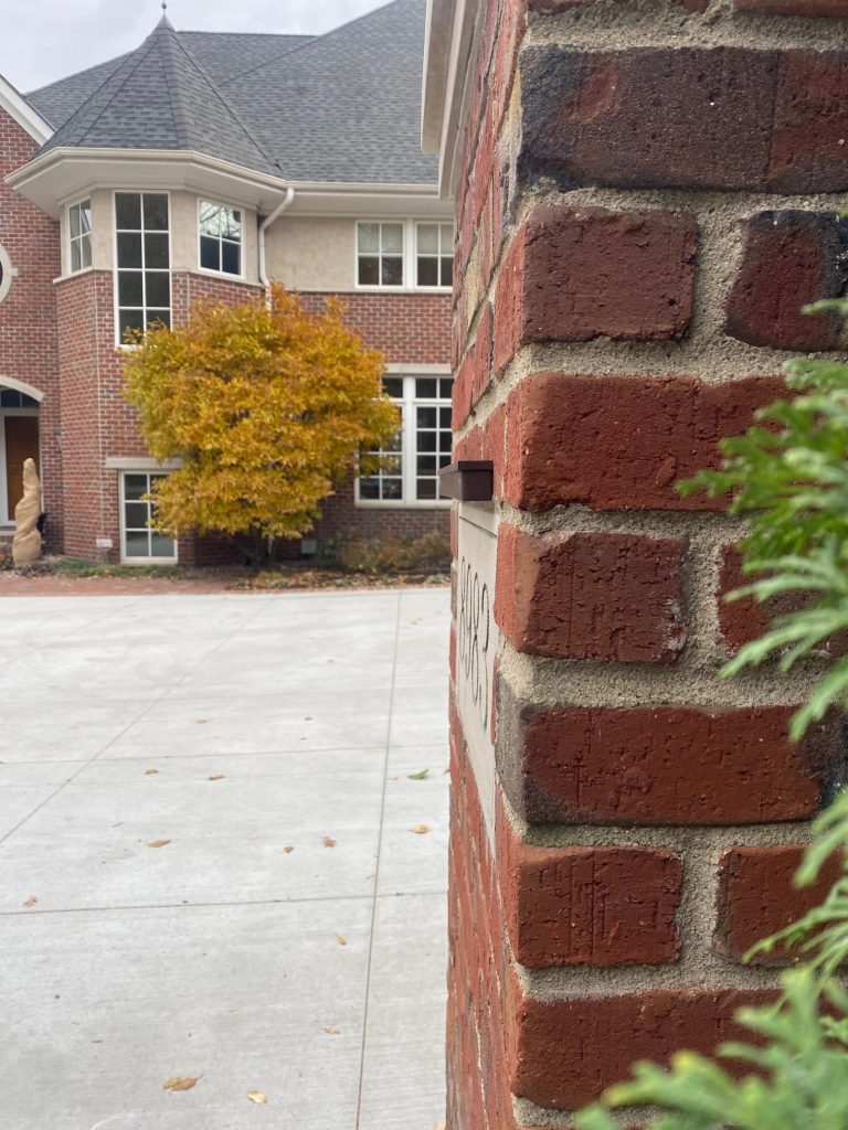 IMG_0837 A brick pillar with a house number partially visible stands in the foreground, featuring a classic Ledge Bar; a large brick house and a yellow-leaved tree are in the background.