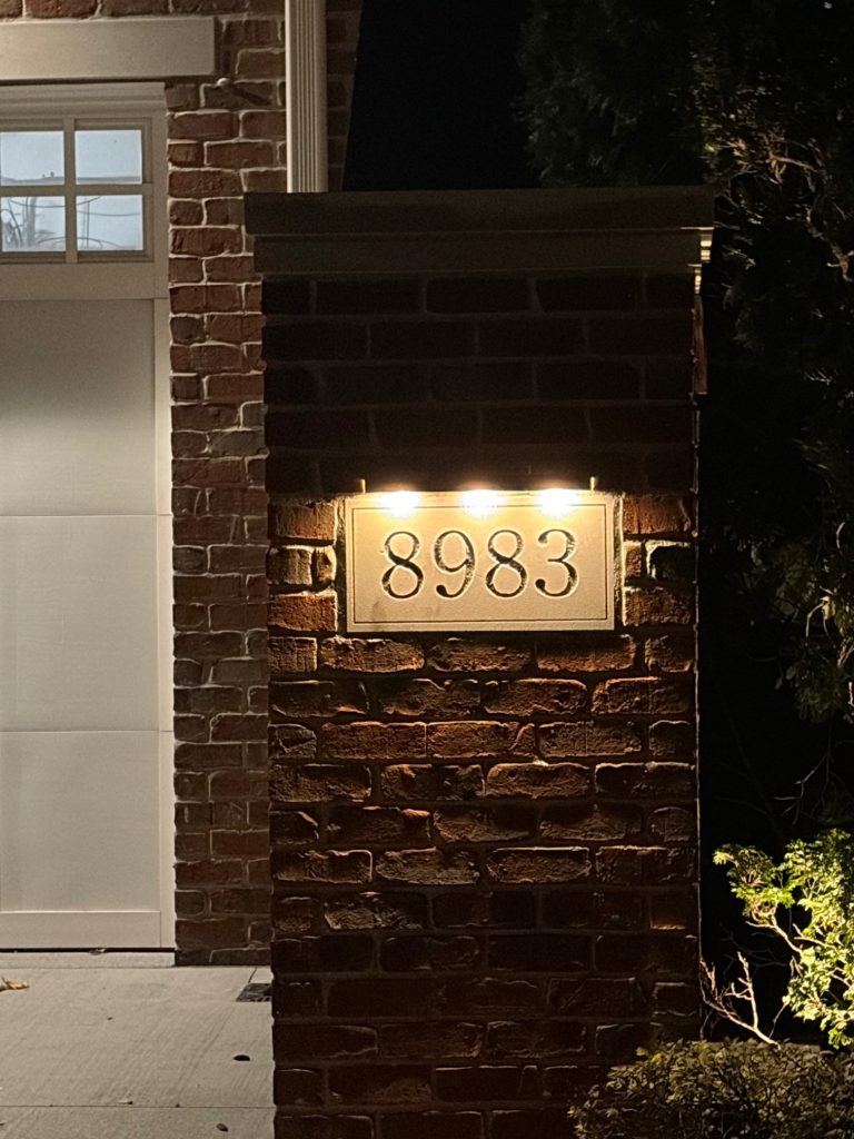 IMG_2616 A brick pillar with the house number 8983 illuminated by three Bar Series lights at night, next to a garage door and some greenery.