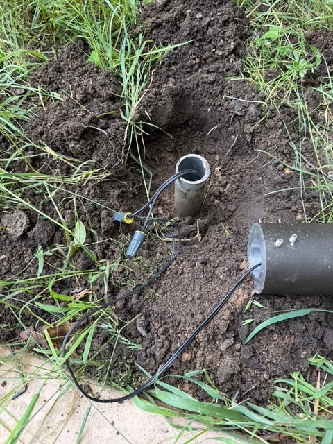 IMG_2636 A cylindrical metal post from the Tubelle Series and wires are seen in a hole dug in soil with grass around, suggesting outdoor electrical or lighting installation or repair work.