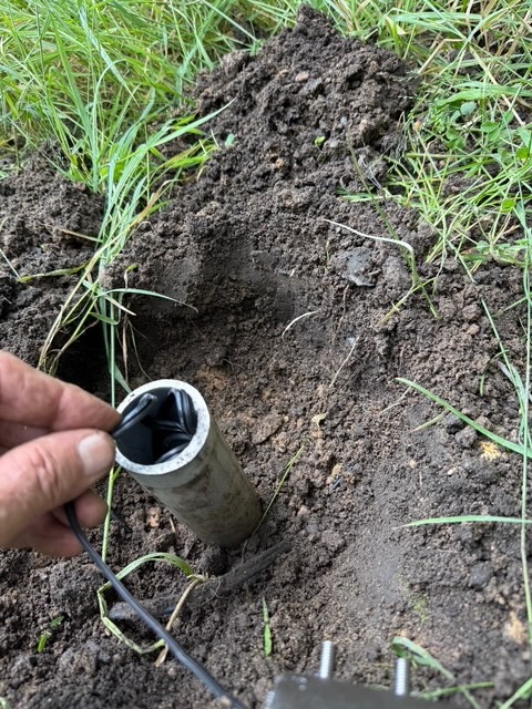 IMG_2637 A hand places a black cable from the Tubelle Series into a metal pipe set in the ground, surrounded by dirt and grass.