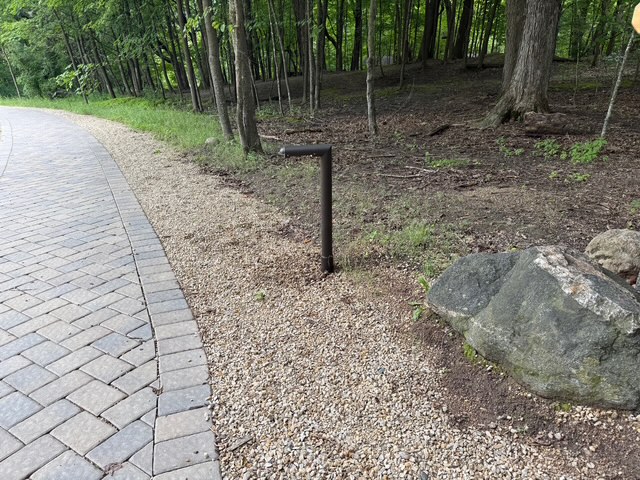 IMG_2645 A Tubelle Series black metal post sticks out of the ground next to a gravel path, near a large rock and bordered by trees in the background.