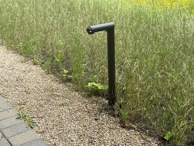 IMG_2650 A black metal Tubelle Series outdoor light fixture stands on a gravel path beside tall grass and wildflowers.