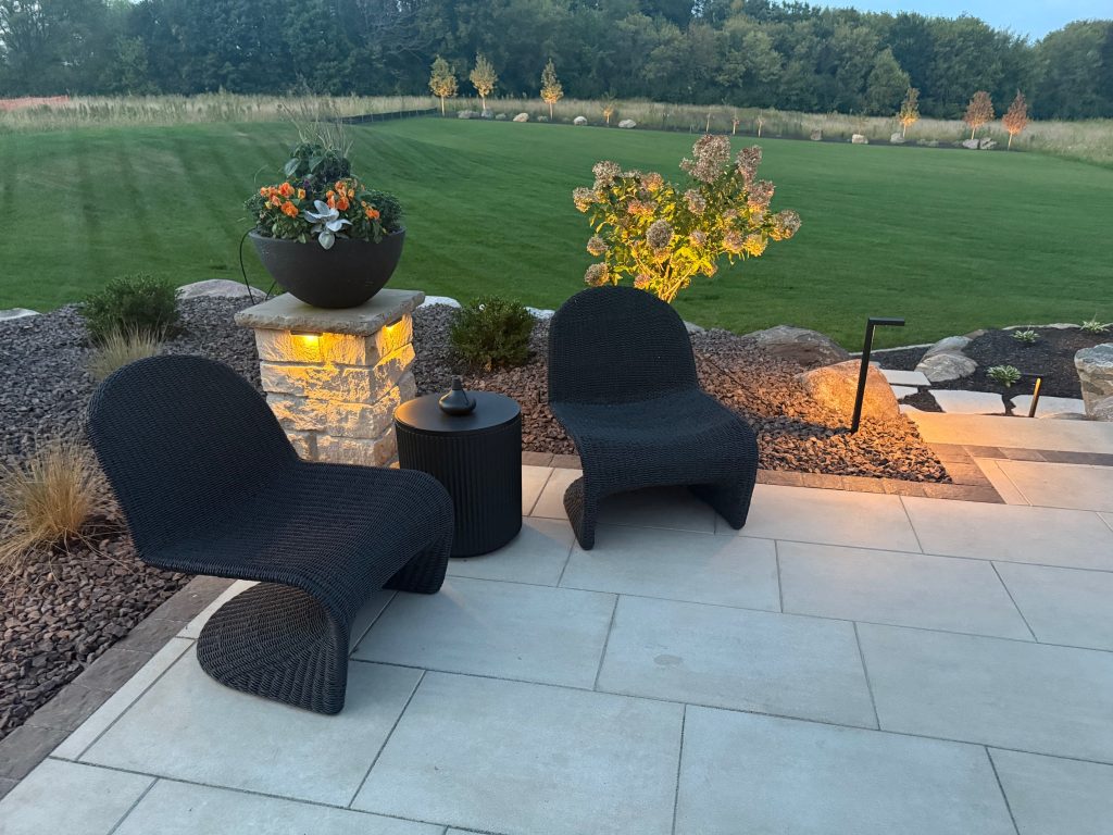 IMG_3073 Two modern black patio chairs from the Ledge Bar Series and a small table are set on a stone terrace overlooking a landscaped yard and green lawn, beautifully highlighted by evening accent lighting.