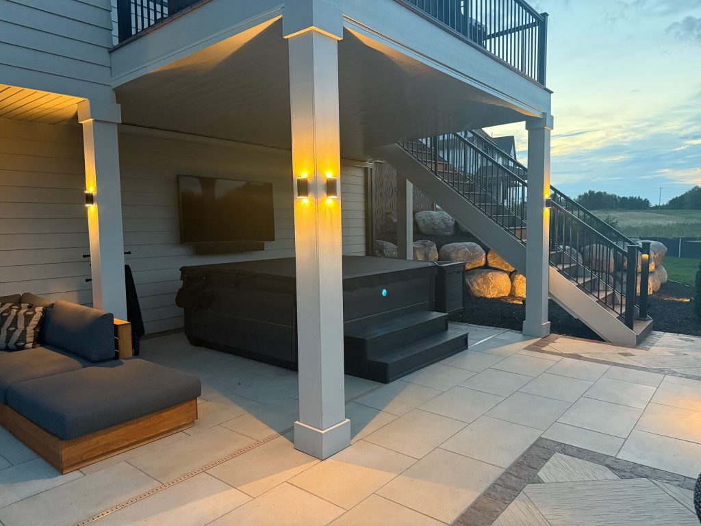 IMG_3077 Outdoor patio area with a hot tub, wall-mounted TV, sectional seating, and stairs leading to an upper deck at dusk. Fence sconce lights are mounted on the pillars for a warm ambiance.