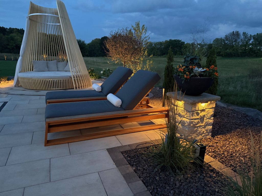IMG_3091 Two wooden lounge chairs with gray cushions and white pillows are on a patio beside a large round daybed, with landscape lighting and greenery in the background at dusk.
