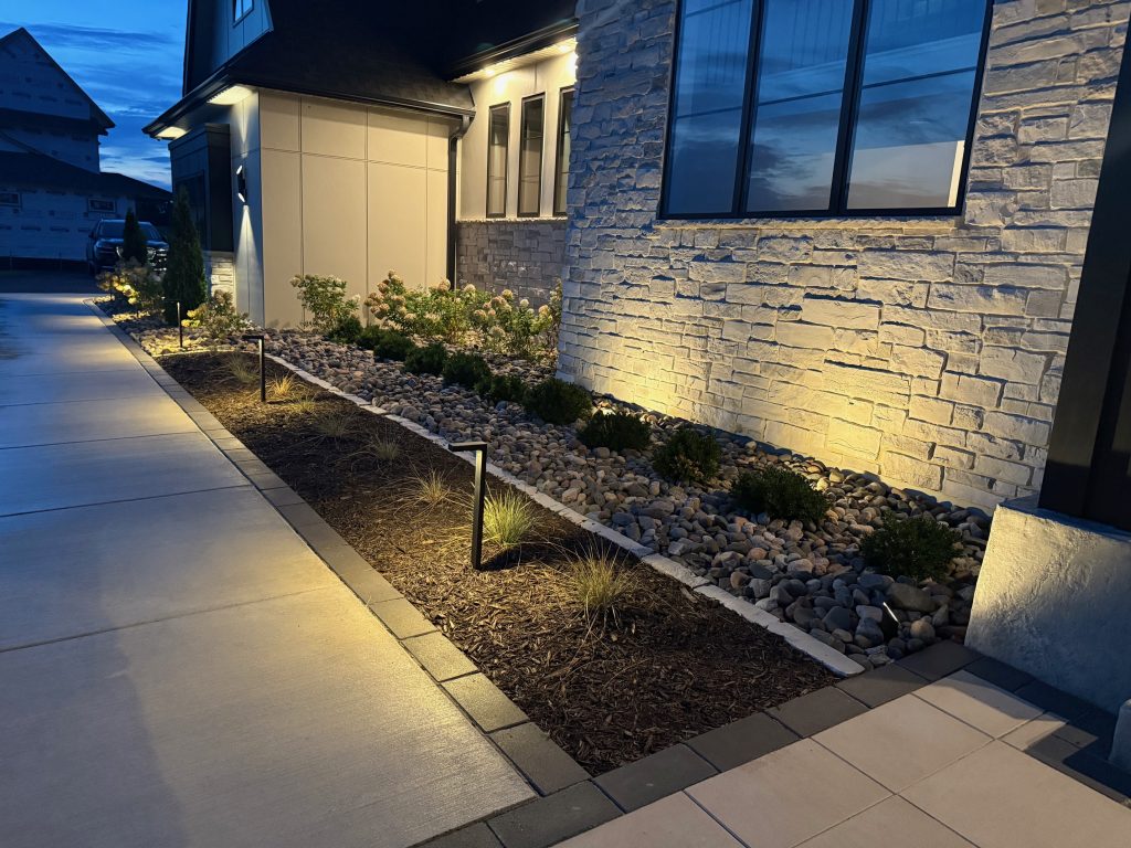 Modern house exterior at dusk, with stone facade, landscape lighting, mulch, river rocks, and neatly trimmed shrubs along a concrete walkway.
