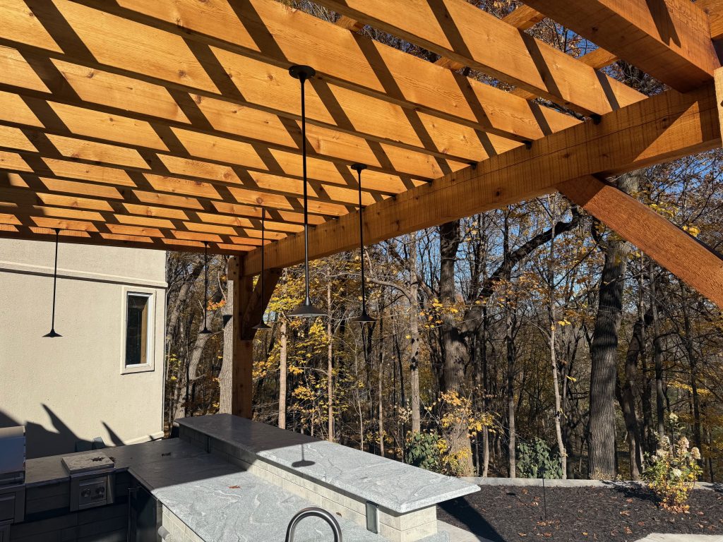 IMG_3432 A wooden pergola provides shade over an outdoor kitchen with a stone countertop and Donovan Series pendant lighting, set against a backdrop of trees with autumn foliage.