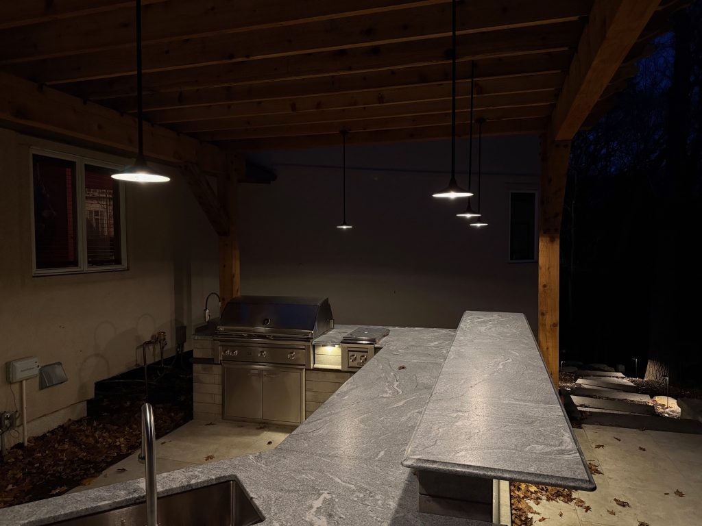 IMG_3484 Outdoor kitchen area under a wooden pergola at night, featuring Donovan Series pendant lamps illuminating the grill, countertops, and sink.