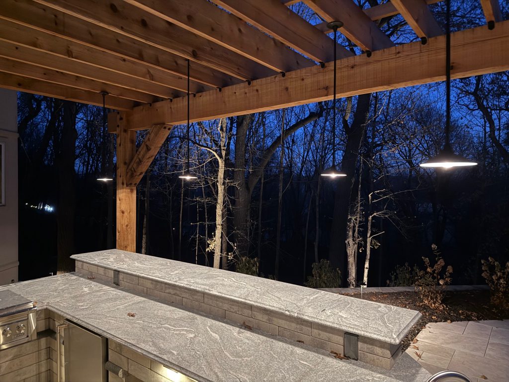 IMG_3485 Outdoor kitchen with a stone countertop and Donovan Series pendant lights, situated under a wooden pergola at dusk with trees in the background.