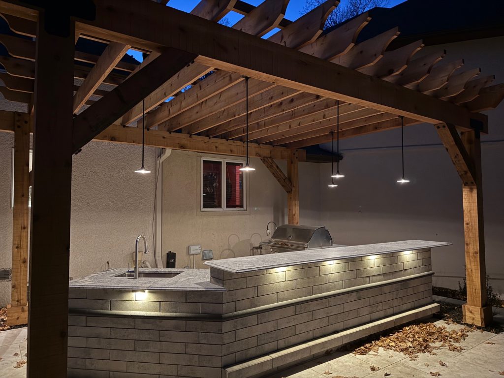 IMG_3489 Outdoor kitchen with a stone counter under a wooden pergola, illuminated by Donovan Series pendant lamps and accent lighting, featuring a built-in grill and sink, photographed at dusk.