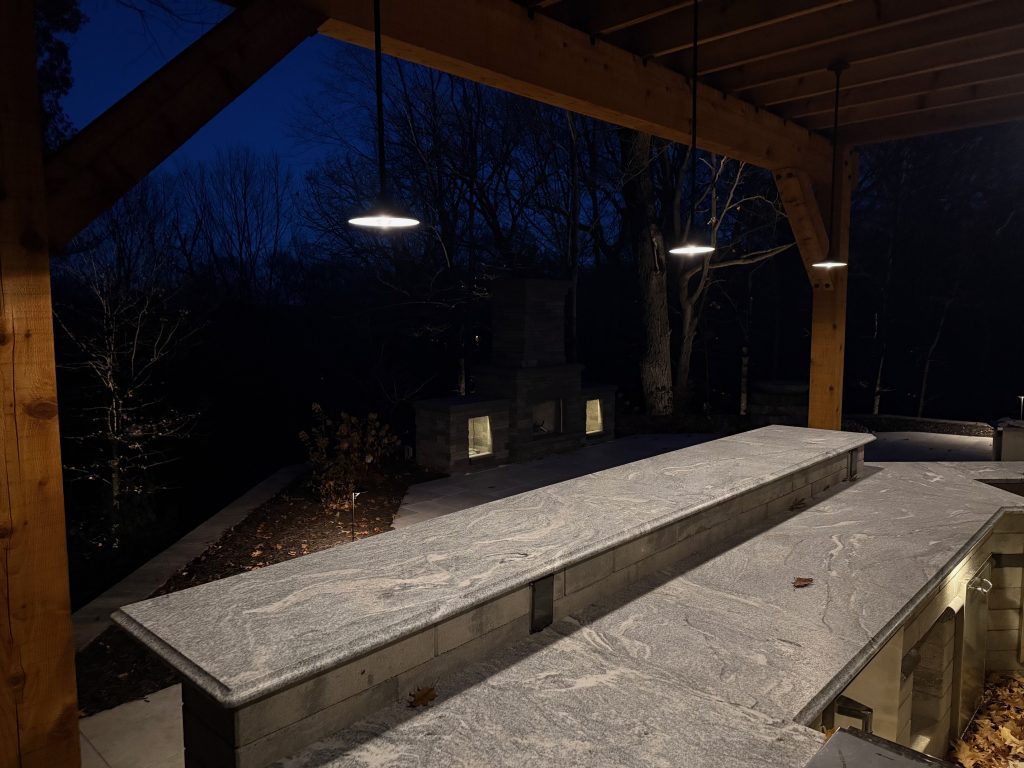 IMG_3491 Outdoor stone bar and countertop under a wooden pergola at night, featuring Donovan Series hanging lights and a lit fireplace in the background.