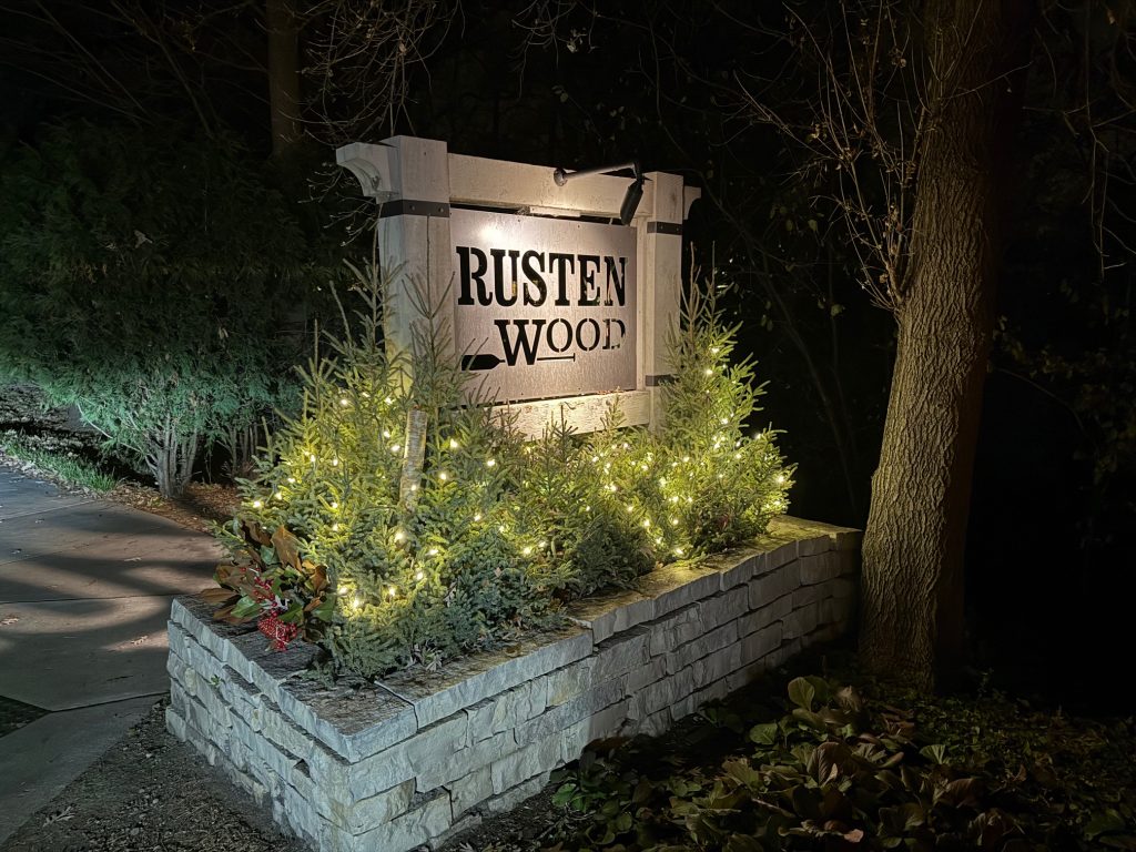 IMG_3556 A stone sign reading "Rusten Wood" is illuminated at night by Brass Series lighting, surrounded by small evergreen bushes decorated with string lights.